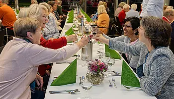 Mehrere Frauen sitzen an einer festlich gedeckten langen Tafel und stoßen mit Weingläsern an.