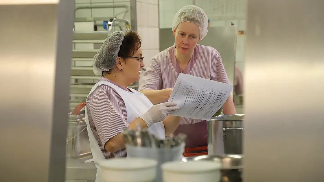 Zwei Mitarbeitende in Arbeitskleidung stehen in einer Großküche und besprechen einen Arbeitsplan anhand von Unterlagen.