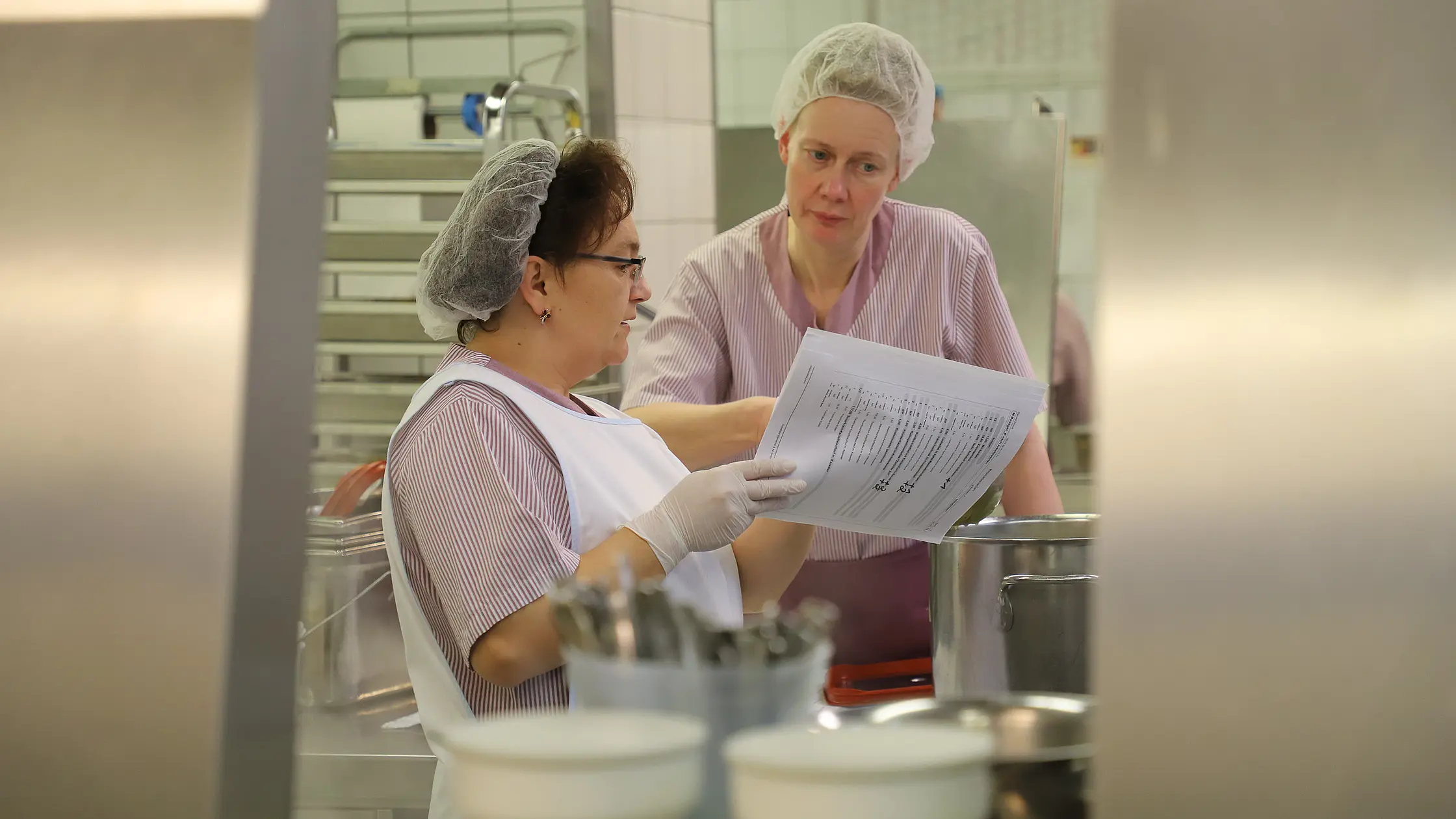 Zwei Mitarbeitende in Arbeitskleidung stehen in einer Großküche und besprechen einen Arbeitsplan anhand von Unterlagen.