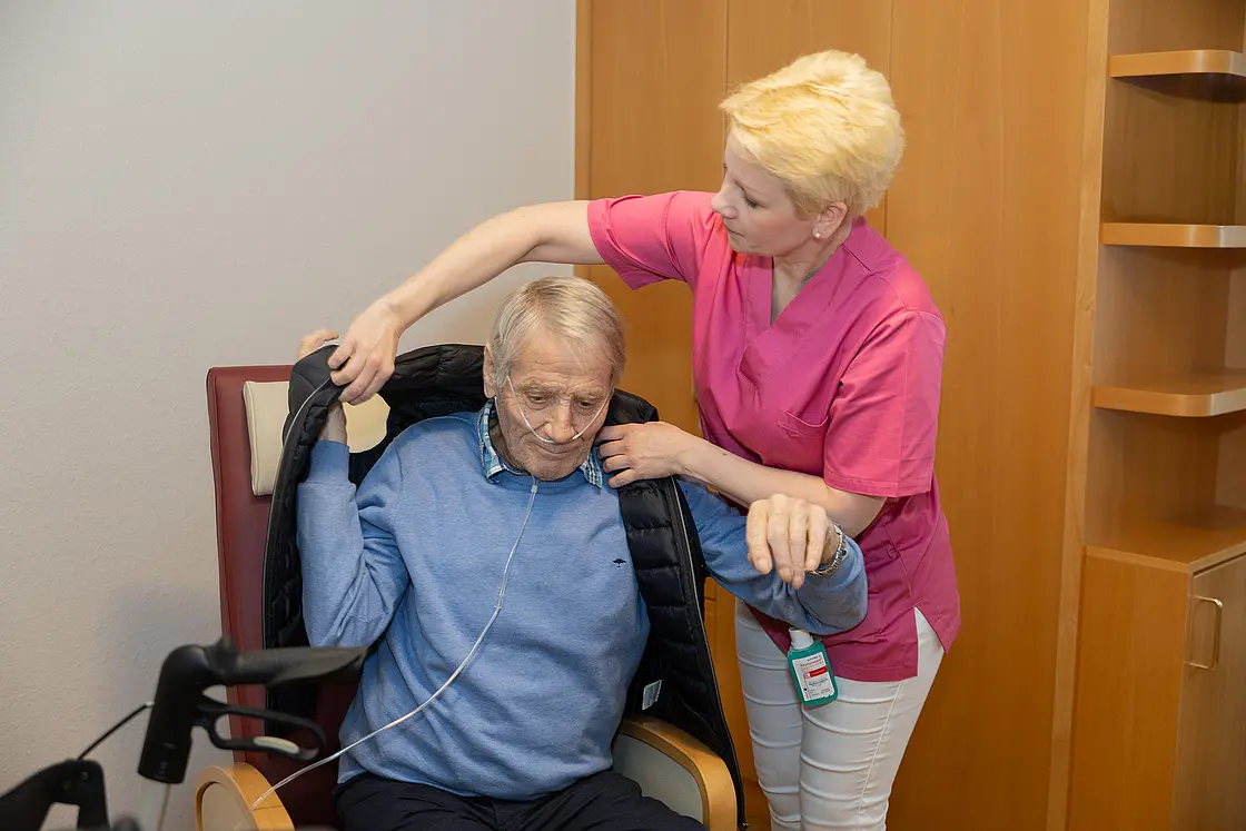 Eine Pflegefachassistentin hilft einem sitzenden Altenheimbewohner beim Anziehen einer Jacke.