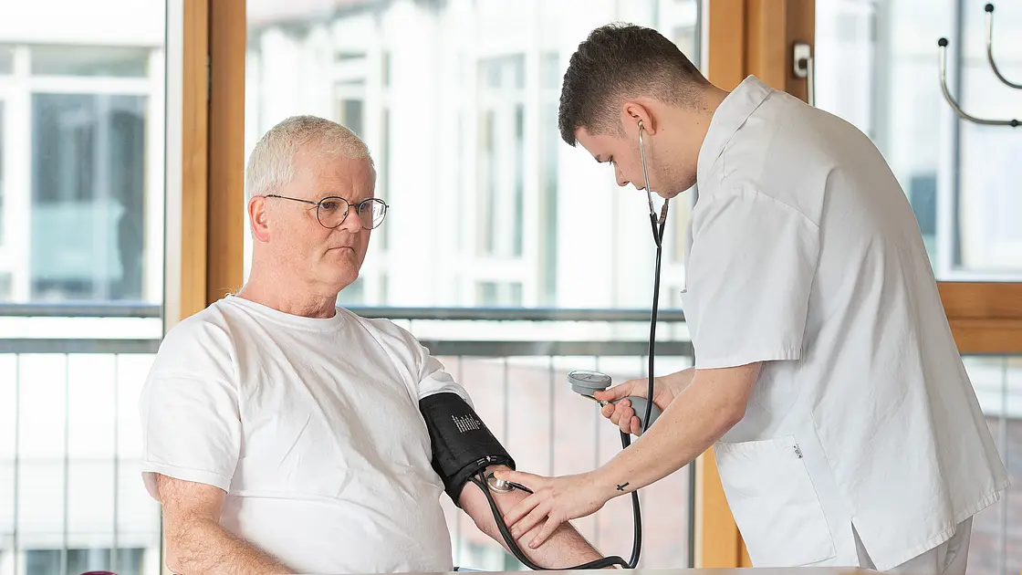 Ein Auszubildender misst einem sitzenden älteren Patienten mit Manschette und Stethoskop den Blutdruck.