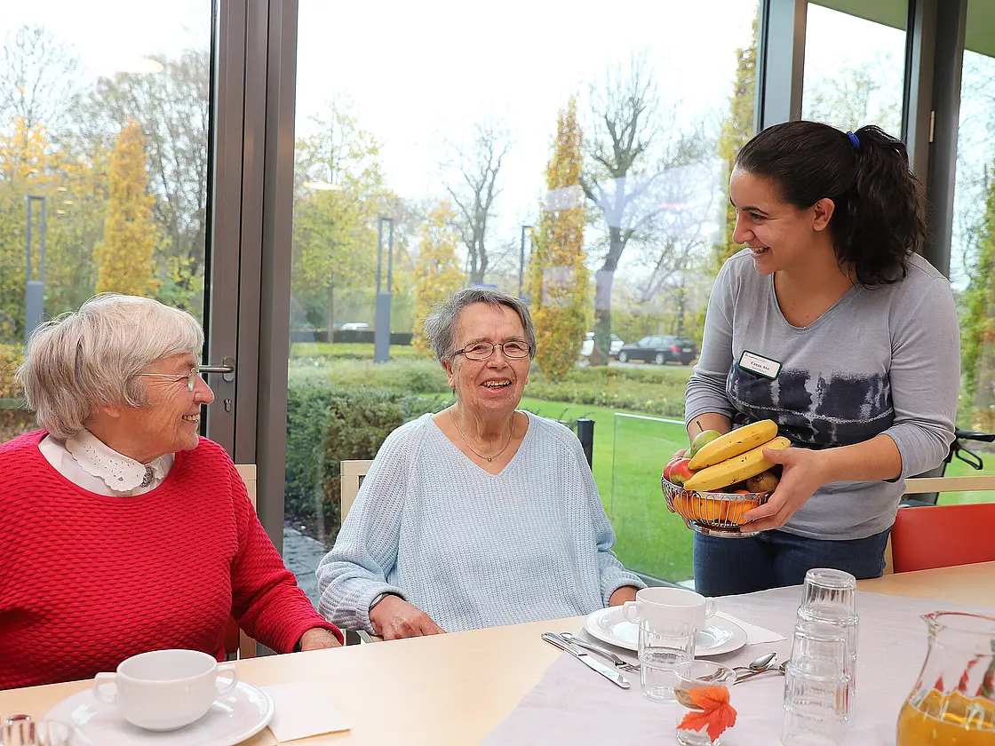 Eine Mitarbeiterin bringt einer Gruppe älterer Frauen frisches Obst an einen gedeckten Tisch.