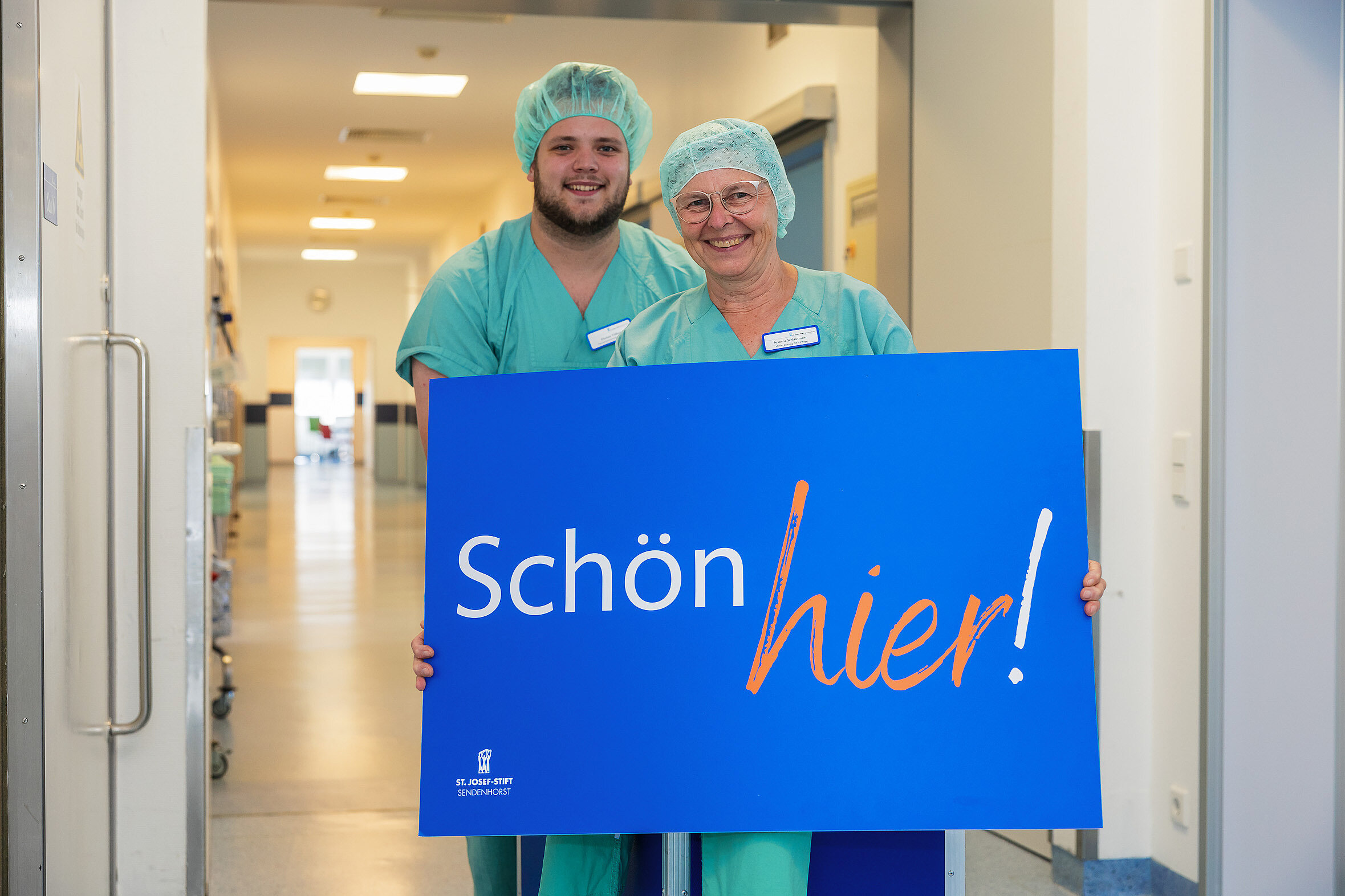 Zwei Mitarbeitende in OP-Kleidung stehen auf einem Flur und halten ein Schild mit der Aufschrift „Schön hier!“.