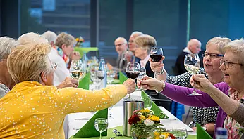 Seniorinnen sitzen an einer langen Tafel und stoßen mit Wein- und Wassergläsern an.