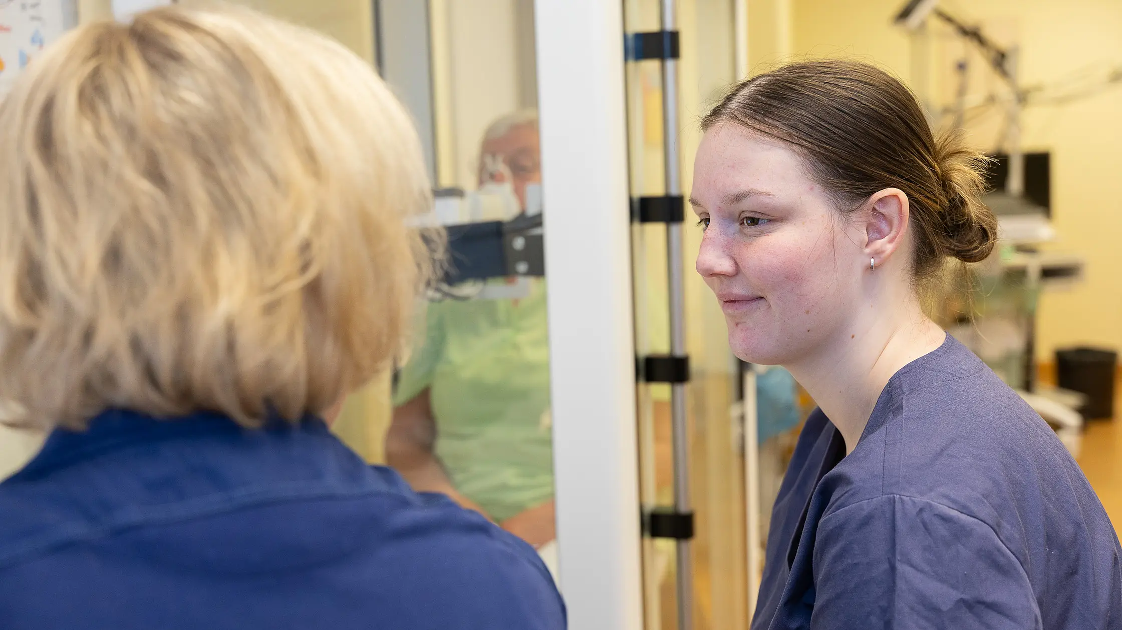 Eine Auszubildende in medizinischer Arbeitskleidung spricht mit einer Kollegin in einem Untersuchungsraum.