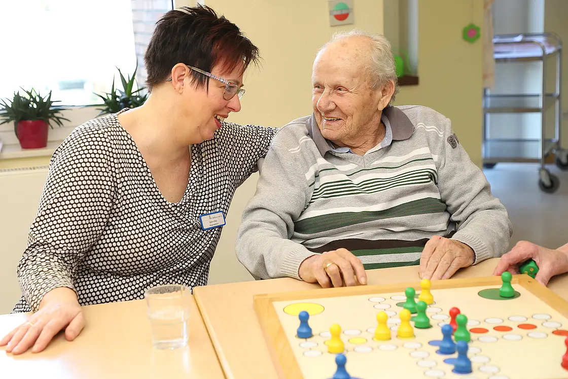 Eine Betreuungshelferin legt lächelnd den Arm um einen älteren Mann, vor ihnen steht das Spiel "Mensch ärgere dich nicht".