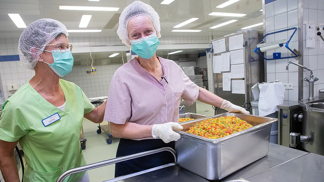 Zwei Mitarbeitende bewegen gemeinsam einen Behälter mit vorbereiteten Speisen durch einen Küchenbereich.