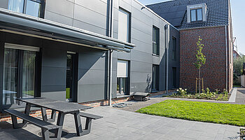 Außenbereich des Apartmenthauses mit Terrasse, Sitzbank und gepflegter Rasenfläche.