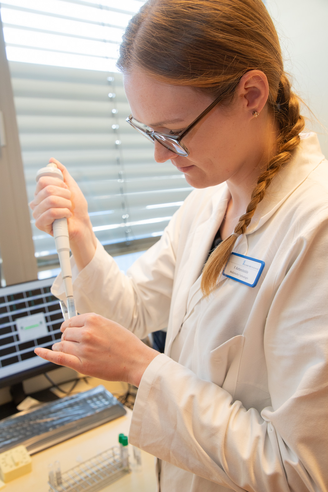 Labor-Mitarbeiterin pipettiert eine Flüssigkeit in ein Probengefäß, im Hintergrund ein Computerarbeitsplatz im Diagnostikzentrum.