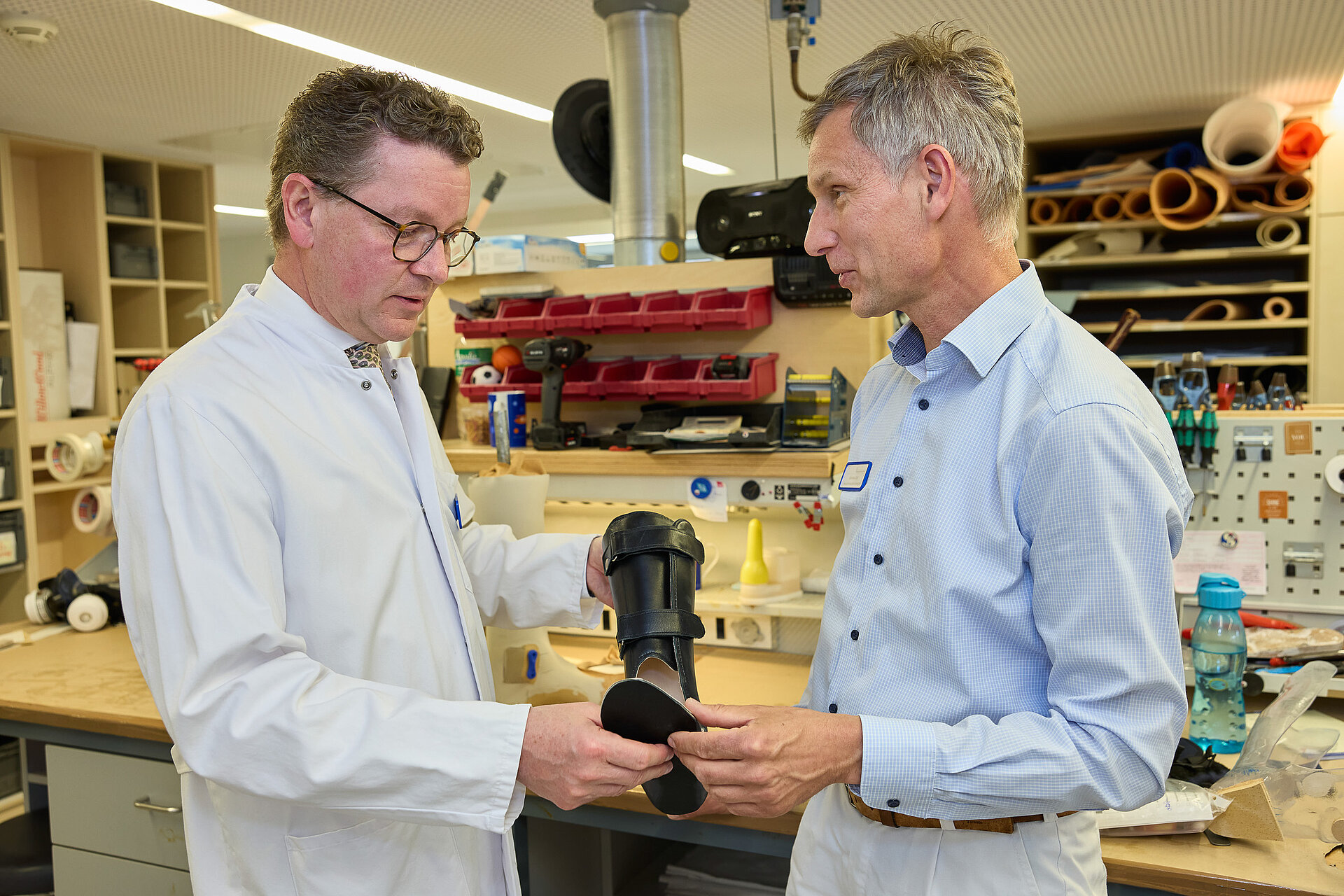Ein Arzt im weißen Kittel bespricht mit einem Mitarbeiter der Orthopädischen Werkstatt eine Orthese in einer Werkstattumgebung.